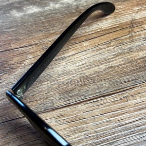 K&ouml;nnte beinhalten: Schwarze Brille mit gl&auml;nzendem Finish, auf einer Holzoberfl&auml;che. Der Rahmen ist abgewinkelt, was das schlanke Design und das Scharnierdetail hervorhebt. Der Text "RAY-BAN" ist auf dem B&uuml;gel sichtbar.