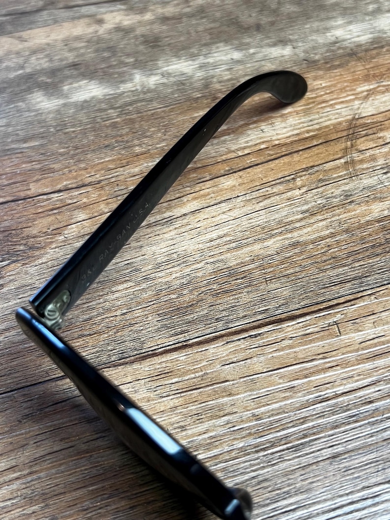 K&ouml;nnte beinhalten: Schwarze Brille mit gl&auml;nzendem Finish, auf einer Holzoberfl&auml;che. Der Rahmen ist abgewinkelt, was das schlanke Design und das Scharnierdetail hervorhebt. Der Text "RAY-BAN" ist auf dem B&uuml;gel sichtbar.
