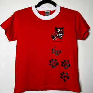 May include: Red t-shirt with a white collar and sleeve trim. The shirt features a tiger graphic and paw prints. The text "Tiger Cubs BSA" is printed below the tiger graphic. The shirt is hanging on a black hanger.