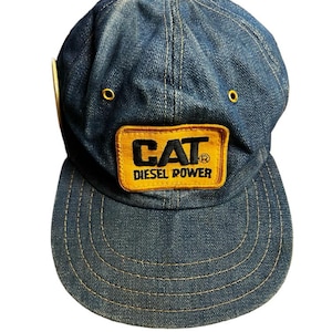 Può includere: Un cappellino da baseball in denim con visiera curva. Il cappellino presenta una toppa gialla con il testo nero "CAT DIESEL POWER". Il cappellino ha cuciture gialle e due occhielli gialli sulla parte superiore.