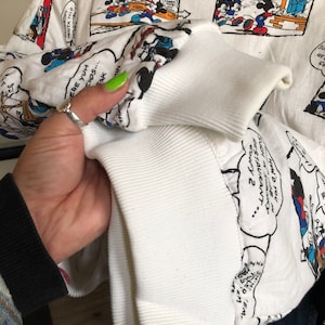 May include: White long-sleeve sweatshirt with a print of Mickey Mouse cartoons. The print features Mickey Mouse and his friends in various scenes. The text "Mickey Mouse" and "Walt Disney's" are printed on the sweatshirt.