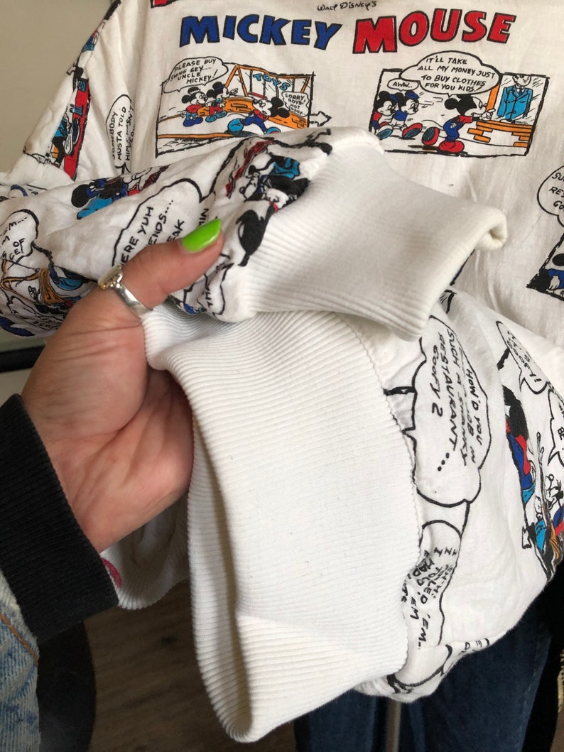 May include: White long-sleeve sweatshirt with a print of Mickey Mouse cartoons. The print features Mickey Mouse and his friends in various scenes. The text "Mickey Mouse" and "Walt Disney's" are printed on the sweatshirt.