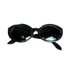 K&ouml;nnte beinhalten: Schwarze ovale Sonnenbrille mit dunklen Gl&auml;sern. Die Sonnenbrille hat einen dicken, abgerundeten Rahmen mit kleinen dekorativen Akzenten auf der Vorderseite. Die B&uuml;gel sind schlank und erstrecken sich nach hinten.