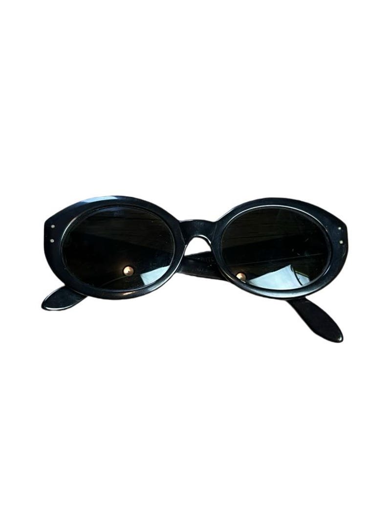 K&ouml;nnte beinhalten: Schwarze ovale Sonnenbrille mit dunklen Gl&auml;sern. Die Sonnenbrille hat einen dicken, abgerundeten Rahmen mit kleinen dekorativen Akzenten auf der Vorderseite. Die B&uuml;gel sind schlank und erstrecken sich nach hinten.