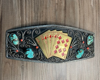 Puede incluir: Una hebilla de cinturón de plata con fondo negro y un diseño de cinco cartas de juego, incluyendo el As, la Reina, el Jack, el 10 y el 9 de diamantes. Las cartas son doradas con diamantes rojos y números rojos. La hebilla está decorada con piedras turquesa y cuentas rojas.