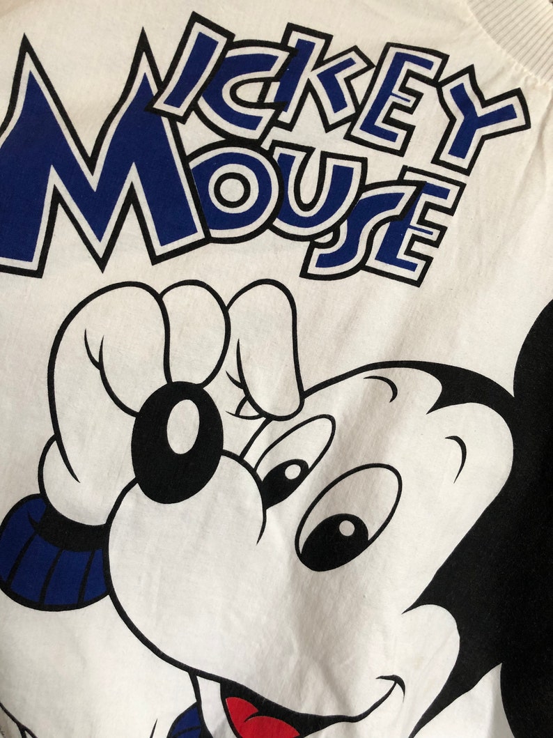 May include: White t-shirt with a black and blue Mickey Mouse graphic. The text "Mickey Mouse" is printed in blue with a black outline.