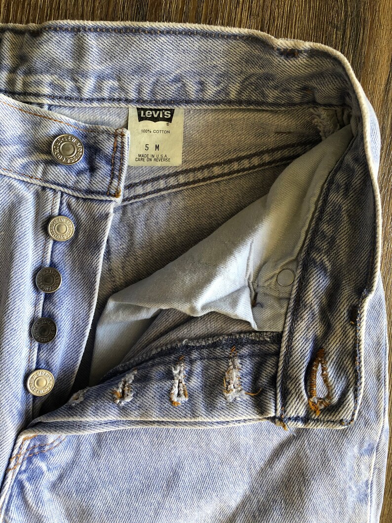Puede incluir: Vaqueros de mezclilla azul claro con una etiqueta Levi's que dice "100% algod&oacute;n" y "5 M" con las palabras "Hecho en EE. UU. Cuidado en el reverso" debajo. Los vaqueros tienen cierre de bot&oacute;n y cinco trabillas para el cintur&oacute;n.