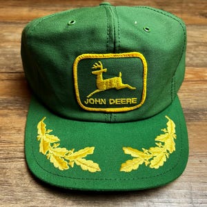 Op de afbeelding: Groene baseballcap met een geel geborduurd John Deere-logo en een geel geborduurd bladerenontwerp op de klep. De cap is gemaakt van een duurzame stof en heeft een klassieke, gestructureerde pasvorm.