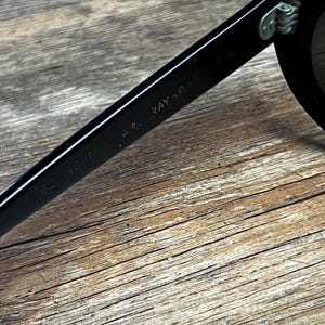 K&ouml;nnte beinhalten: Nahaufnahme einer schwarzen Sonnenbrille mit dem Text "RAY-BAN U.S.A." auf dem B&uuml;gel. Die Sonnenbrille liegt auf einer Holzoberfl&auml;che.