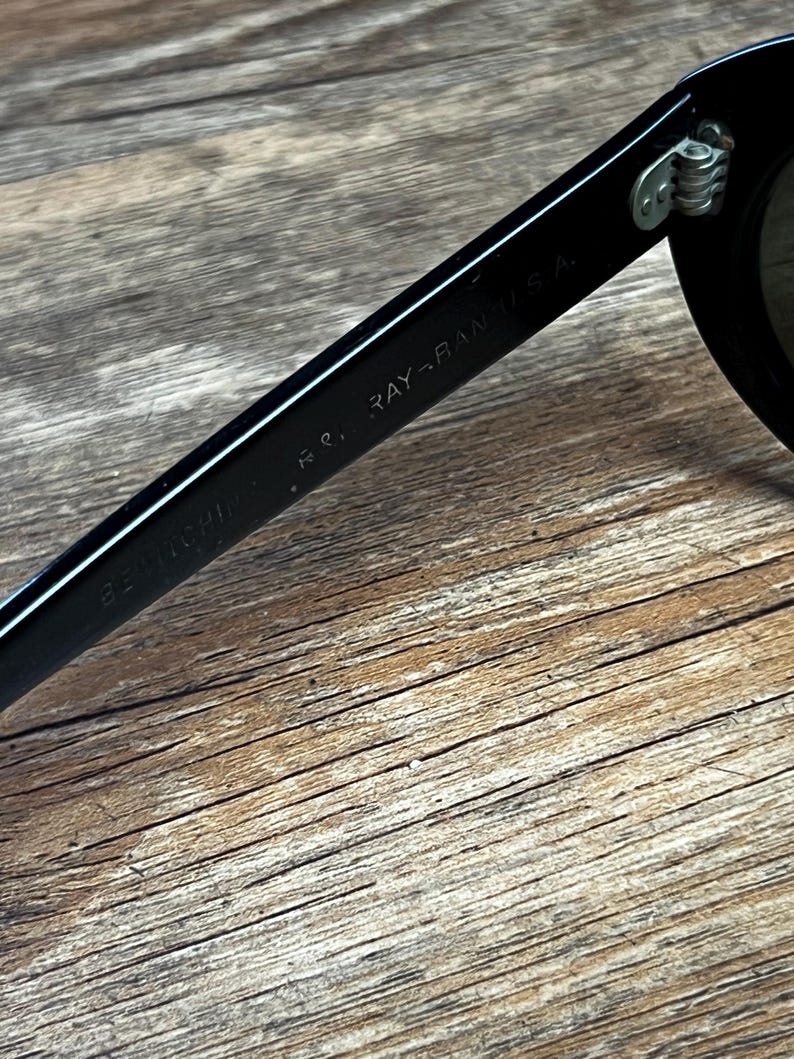 K&ouml;nnte beinhalten: Nahaufnahme einer schwarzen Sonnenbrille mit dem Text "RAY-BAN U.S.A." auf dem B&uuml;gel. Die Sonnenbrille liegt auf einer Holzoberfl&auml;che.