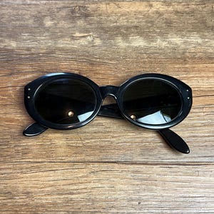 K&ouml;nnte beinhalten: Schwarze, ovale Sonnenbrille mit dunklen Gl&auml;sern. Die Sonnenbrille hat einen dicken Rahmen und kleine dekorative Details an den B&uuml;geln. Sie liegt auf einer Holzoberfl&auml;che.