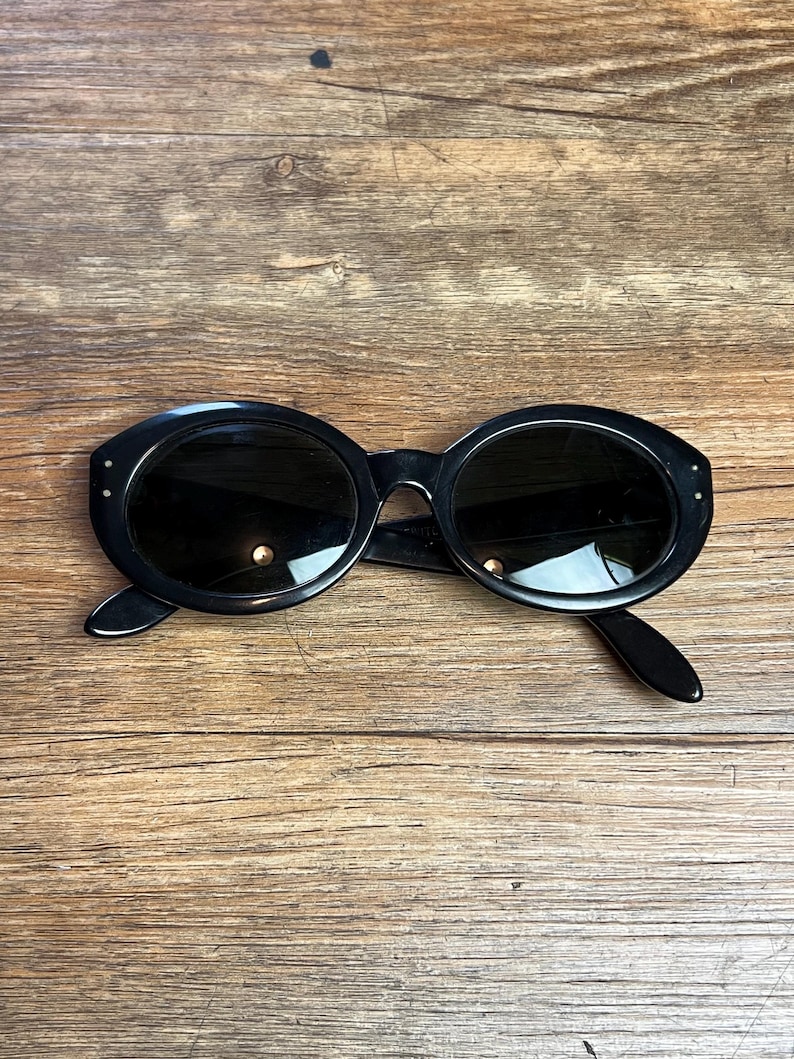 K&ouml;nnte beinhalten: Schwarze, ovale Sonnenbrille mit dunklen Gl&auml;sern. Die Sonnenbrille hat einen dicken Rahmen und kleine dekorative Details an den B&uuml;geln. Sie liegt auf einer Holzoberfl&auml;che.