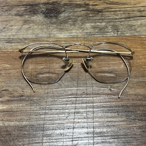 Puede incluir: Gafas con montura dorada con un diseño sin montura. Las gafas tienen una montura fina y delicada y un doble puente. Las lentes son transparentes y las almohadillas nasales son de color oro claro. Las gafas están sobre un fondo de madera.