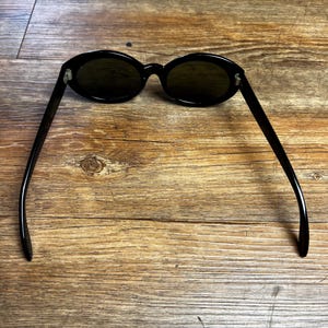 K&ouml;nnte beinhalten: Schwarze ovale Sonnenbrille mit dunklen Gl&auml;sern. Die Sonnenbrille hat ein schlankes, modernes Design mit einem d&uuml;nnen Rahmen und B&uuml;geln. Die Gl&auml;ser sind dunkelgr&uuml;n. Die Sonnenbrille liegt auf einem Holzgrund.