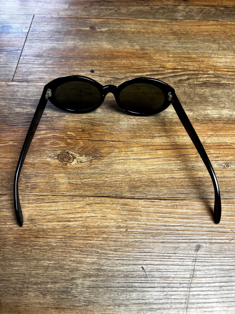 K&ouml;nnte beinhalten: Schwarze ovale Sonnenbrille mit dunklen Gl&auml;sern. Die Sonnenbrille hat ein schlankes, modernes Design mit einem d&uuml;nnen Rahmen und B&uuml;geln. Die Gl&auml;ser sind dunkelgr&uuml;n. Die Sonnenbrille liegt auf einem Holzgrund.
