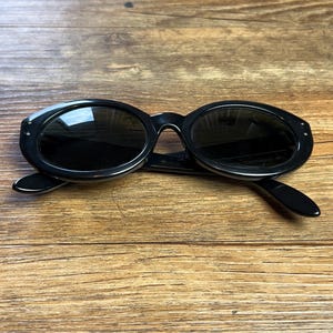 K&ouml;nnte beinhalten: Schwarze ovale Sonnenbrille mit dunklen Gl&auml;sern. Die Sonnenbrille hat einen dicken Rahmen und liegt auf einem Holzgrund. Die B&uuml;gel der Sonnenbrille sind ausgeklappt.