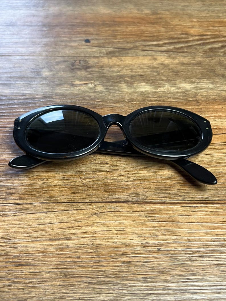 K&ouml;nnte beinhalten: Schwarze ovale Sonnenbrille mit dunklen Gl&auml;sern. Die Sonnenbrille hat einen dicken Rahmen und liegt auf einem Holzgrund. Die B&uuml;gel der Sonnenbrille sind ausgeklappt.