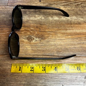 K&ouml;nnte beinhalten: Schwarze Sonnenbrille mit dunklen Gl&auml;sern, ca. 15 cm lang, wird auf einer Holzoberfl&auml;che neben einem gelben Ma&szlig;band gezeigt. Die Sonnenbrille hat ein klassisches Design mit einem schlanken, modernen Look.