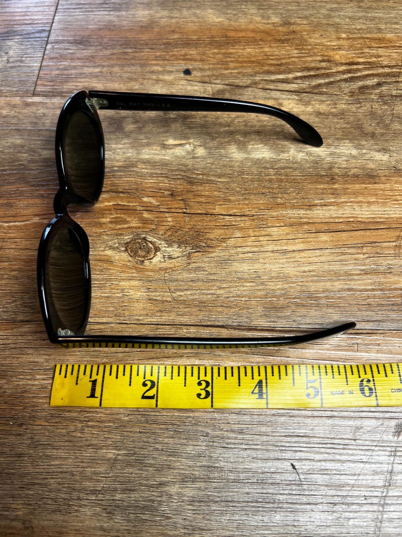 K&ouml;nnte beinhalten: Schwarze Sonnenbrille mit dunklen Gl&auml;sern, ca. 15 cm lang, wird auf einer Holzoberfl&auml;che neben einem gelben Ma&szlig;band gezeigt. Die Sonnenbrille hat ein klassisches Design mit einem schlanken, modernen Look.