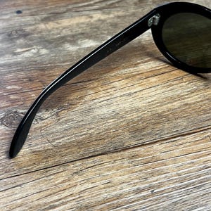 K&ouml;nnte beinhalten: Schwarze Sonnenbrille mit runden Gl&auml;sern und einem dicken Rahmen. Die Sonnenbrille hat ein klassisches Design und ist vor einem Holzgrund platziert. Der Text "RAY-BAN" ist auf dem B&uuml;gel der Sonnenbrille zu sehen.