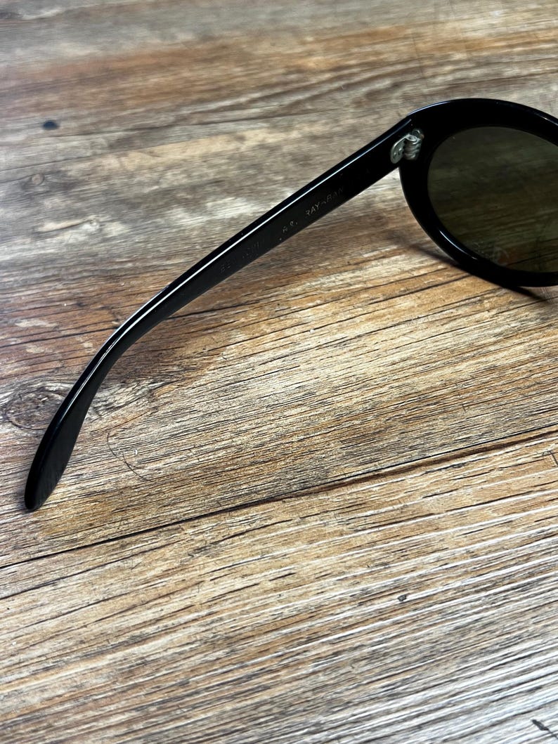 K&ouml;nnte beinhalten: Schwarze Sonnenbrille mit runden Gl&auml;sern und einem dicken Rahmen. Die Sonnenbrille hat ein klassisches Design und ist vor einem Holzgrund platziert. Der Text "RAY-BAN" ist auf dem B&uuml;gel der Sonnenbrille zu sehen.
