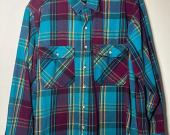 Può includere: Una camicia a quadri a maniche lunghe in verde acqua, viola e giallo. La camicia ha due tasche frontali con chiusura a bottone e un'abbottonatura frontale. Il colletto e i polsini sono verde acqua. La camicia è appesa a una gruccia nera.