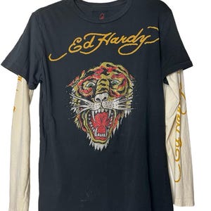 May include: Black t-shirt with a tiger graphic and the text "Ed Hardy" in gold. The shirt has long, cream-colored sleeves with gold text. The tiger graphic is detailed with red, yellow, and green accents.