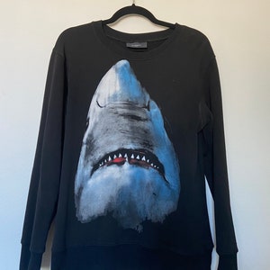 Pode incluir: Camisola de moletom preta com uma grande estampa gráfica de uma cabeça de tubarão em tons de azul e cinza.