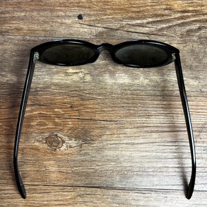 K&ouml;nnte beinhalten: Schwarze, ovale Sonnenbrille mit dunklen Gl&auml;sern. Die Sonnenbrille hat ein schlankes Design mit d&uuml;nnen, geraden B&uuml;geln. Der Rahmen und die B&uuml;gel sind gl&auml;nzend schwarz, und die Gl&auml;ser sind dunkel und bieten Schatten. Die Sonnenbrille liegt auf einem Holzgrund.