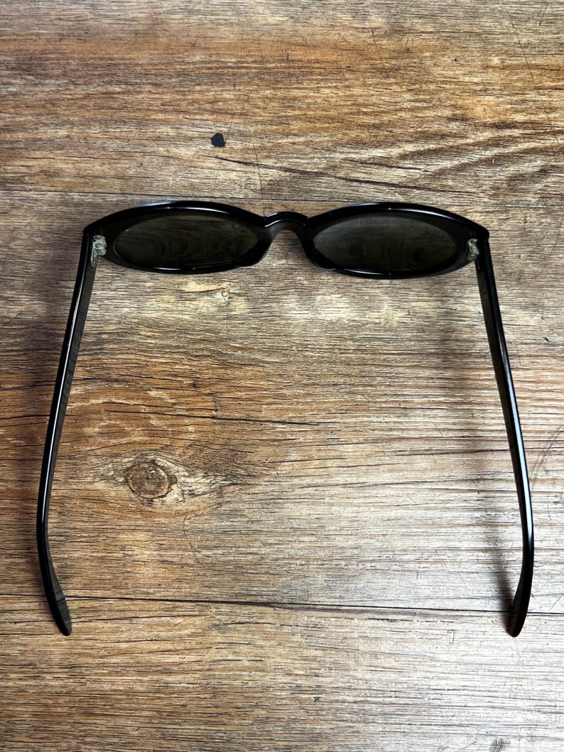 K&ouml;nnte beinhalten: Schwarze, ovale Sonnenbrille mit dunklen Gl&auml;sern. Die Sonnenbrille hat ein schlankes Design mit d&uuml;nnen, geraden B&uuml;geln. Der Rahmen und die B&uuml;gel sind gl&auml;nzend schwarz, und die Gl&auml;ser sind dunkel und bieten Schatten. Die Sonnenbrille liegt auf einem Holzgrund.