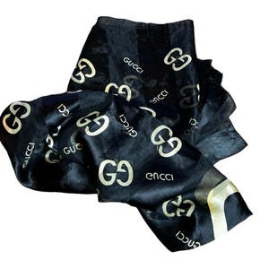 May include: Black scarf with a repeating white Gucci logo pattern. The scarf is made of a silky material and is folded and draped. The Gucci logo is visible throughout the scarf.