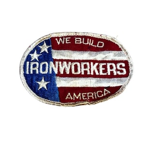 May include: An embroidered patch in an oval shape, featuring the American flag design. The patch has a blue field with white stars and red and white stripes. The words "WE BUILD IRONWORKERS AMERICA" are embroidered in white.