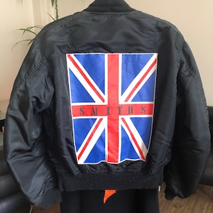 Puede incluir: Una chaqueta bomber negra con una bandera del Reino Unido y la palabra "SMITHS" impresa en la espalda.