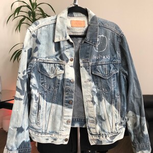 May include: A light blue denim jacket with a white bleach-splattered design. The jacket has a white embroidered design on the back that says "Bauhaus" and a circle with a line through it.