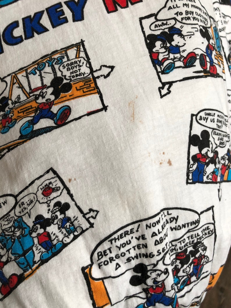 May include: White t-shirt with a repeating pattern of Mickey Mouse cartoon illustrations. The illustrations feature Mickey Mouse in various poses and settings, including playing with toys, riding a swing set, and interacting with other characters. The text "Mickey Mouse" is printed in blue at the top of the shirt.
