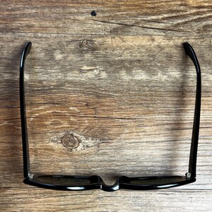 K&ouml;nnte beinhalten: Schwarze Sonnenbrille mit klassischem rechteckigem Rahmen. Die Sonnenbrille hat dunkle Gl&auml;ser und geschwungene B&uuml;gel. Das Bild zeigt die Sonnenbrille auf einer Holzoberfl&auml;che, die ihre Form und ihren Stil hervorhebt.