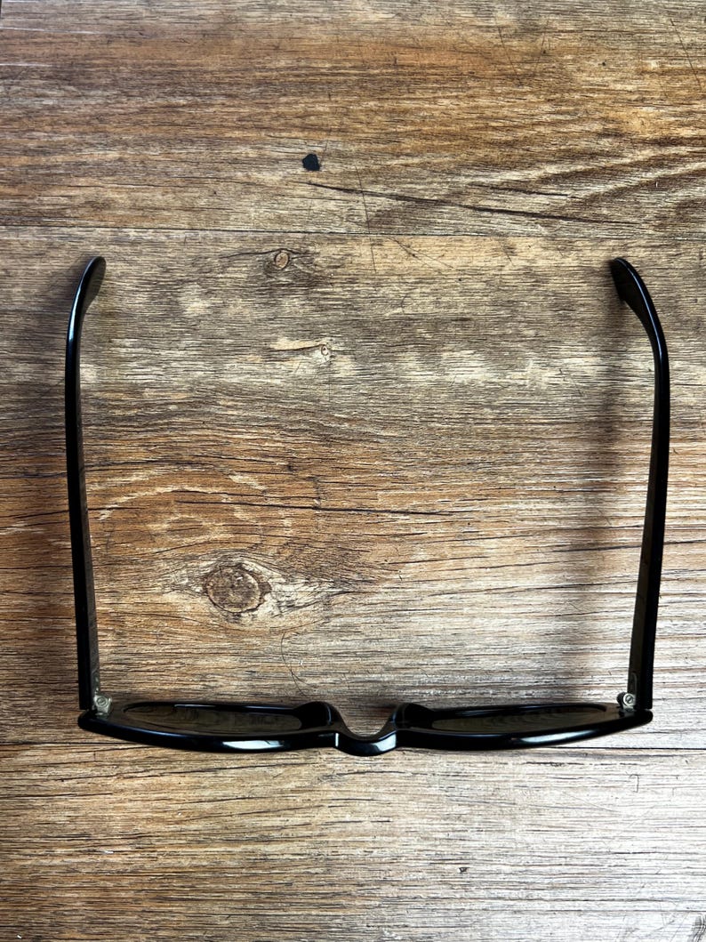 K&ouml;nnte beinhalten: Schwarze Sonnenbrille mit klassischem rechteckigem Rahmen. Die Sonnenbrille hat dunkle Gl&auml;ser und geschwungene B&uuml;gel. Das Bild zeigt die Sonnenbrille auf einer Holzoberfl&auml;che, die ihre Form und ihren Stil hervorhebt.