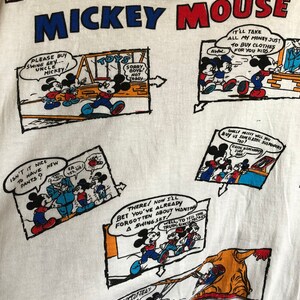 May include: White t-shirt with a repeating pattern of Mickey Mouse comic strips. The shirt features Mickey Mouse in various scenes, including him buying clothes, playing on a swing set, and wearing new pants. The text on the shirt reads "Walt Disney's Mickey Mouse".