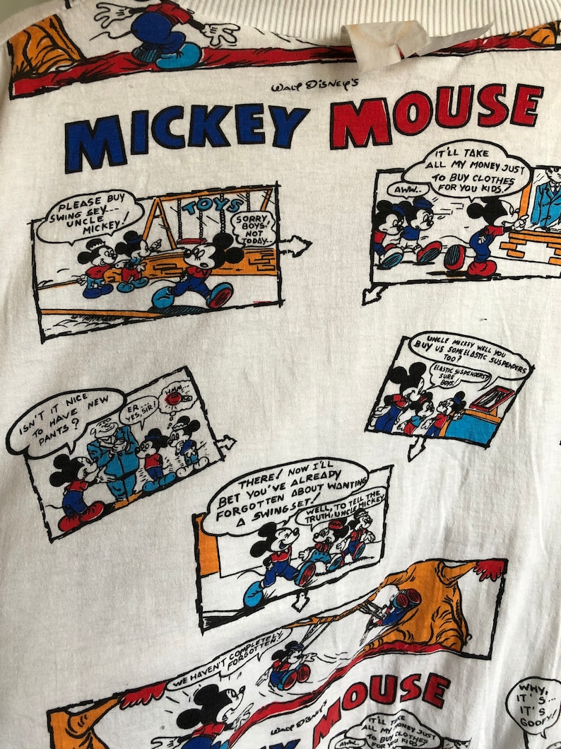 May include: White t-shirt with a repeating pattern of Mickey Mouse comic strips. The shirt features Mickey Mouse in various scenes, including him buying clothes, playing on a swing set, and wearing new pants. The text on the shirt reads "Walt Disney's Mickey Mouse".