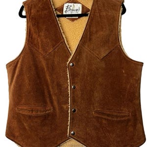 Può includere: Un gilet in pelle scamosciata marrone con fodera in sherpa beige e due tasche frontali. Il gilet ha una chiusura con bottoni e un'etichetta con la scritta "Berman's". Il gilet è appeso a una gruccia nera.
