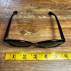 K&ouml;nnte beinhalten: Schwarze Cat-Eye-Sonnenbrille mit gelbem Ma&szlig;band. Die Sonnenbrille hat ein klassisches Retro-Design mit schmalem Rahmen. Das Ma&szlig;band zeigt Messungen von 2,5 bis 15 cm an.