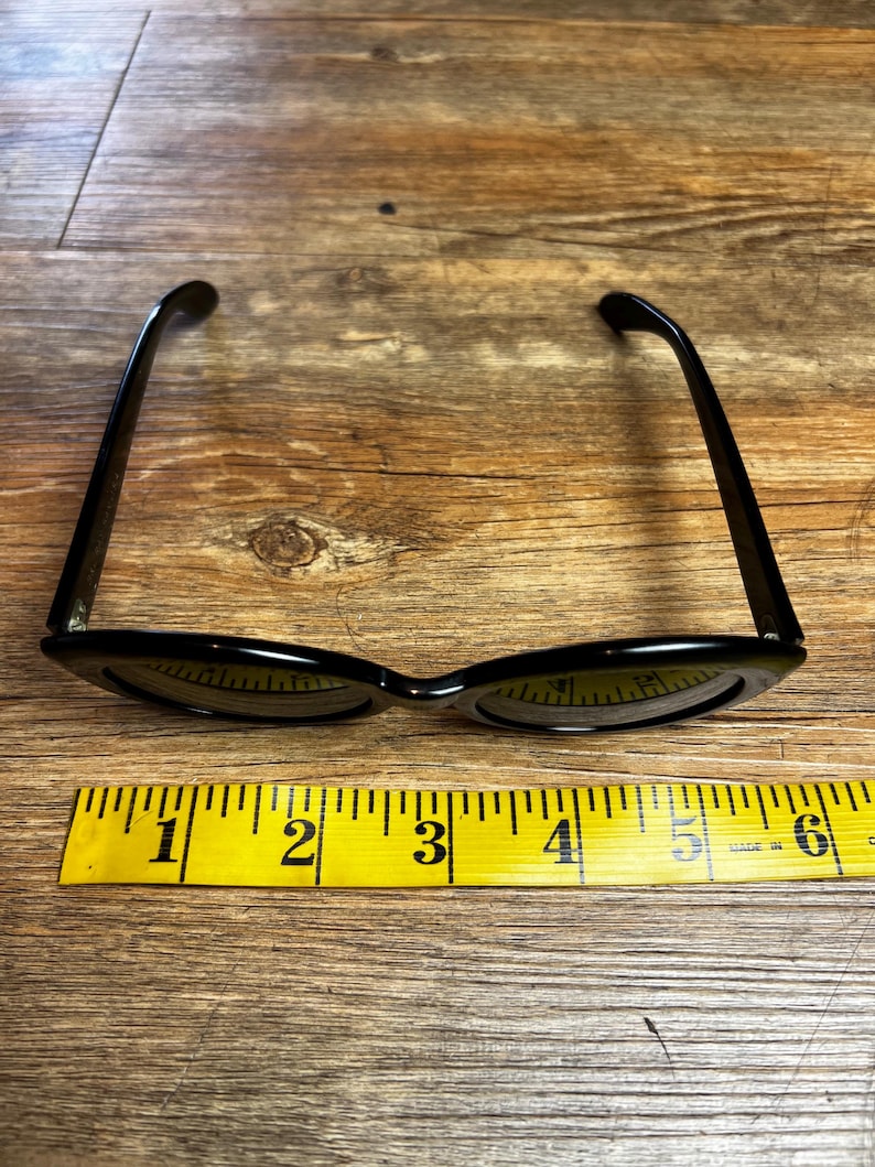 K&ouml;nnte beinhalten: Schwarze Cat-Eye-Sonnenbrille mit gelbem Ma&szlig;band. Die Sonnenbrille hat ein klassisches Retro-Design mit schmalem Rahmen. Das Ma&szlig;band zeigt Messungen von 2,5 bis 15 cm an.