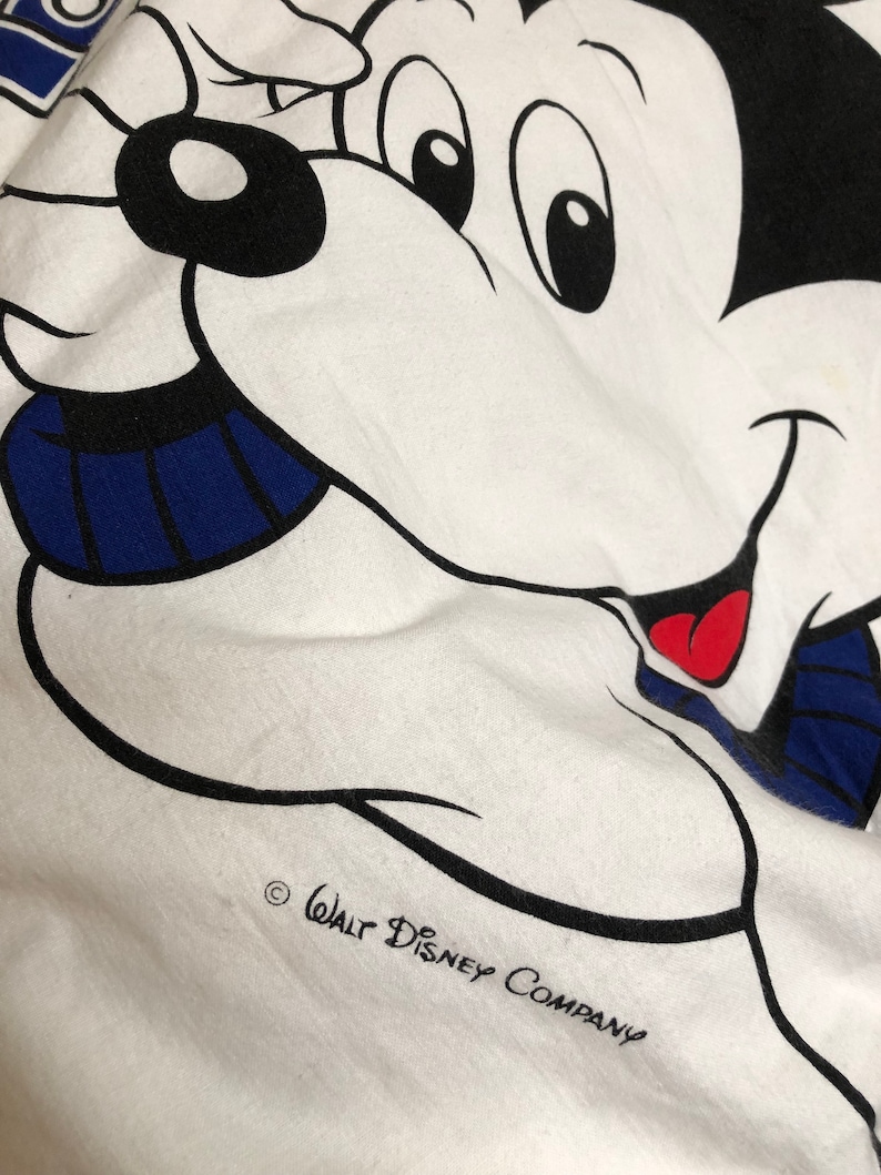 May include: White t-shirt with a cartoon image of Mickey Mouse wearing a blue and white striped scarf. The text "Walt Disney Company" is printed in black at the bottom of the shirt.