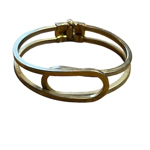 May include: A gold-toned metal hinged bangle bracelet featuring a double-band design that converges into a central oval loop. The bracelet has a smooth, polished finish and a secure mechanical clasp mechanism at the top for opening and closing.