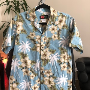 Op de afbeelding: Een lichtblauw, kort mouwig overhemd met knopen en een tropische print met palmbomen en witte bloemen. Het overhemd heeft een knoopsluiting en een zakje aan de linkerkant. Het label op het overhemd luidt "Reyn Spooner Hawaii".