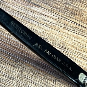 K&ouml;nnte beinhalten: Nahaufnahme eines schwarzen Ray-Ban-Brillenrahmens mit dem Text "BEWITCHING" und "H&L RAY-BAN U.S.A." auf dem B&uuml;gel. Der Rahmen liegt auf einer Holzoberfl&auml;che.