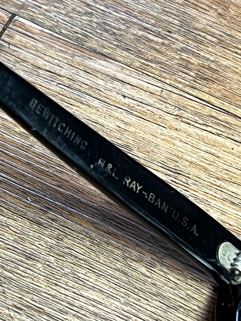 K&ouml;nnte beinhalten: Nahaufnahme eines schwarzen Ray-Ban-Brillenrahmens mit dem Text "BEWITCHING" und "H&L RAY-BAN U.S.A." auf dem B&uuml;gel. Der Rahmen liegt auf einer Holzoberfl&auml;che.