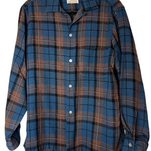 May include: A long-sleeve button-down shirt featuring a plaid pattern in shades of blue, brown, and black. The shirt has a collared neckline, a single chest pocket, and white buttons. An Arrow brand label is visible on the inside collar.