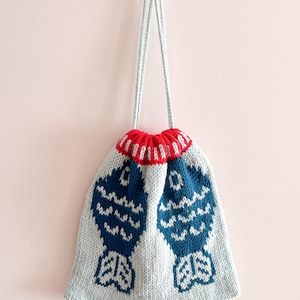Puede incluir: Un bolso de punto con cordón. El bolso tiene un cuerpo azul claro y una parte superior a rayas rojas y blancas. Dos grandes diseños de peces azules decoran el bolso. El cordón es azul claro.