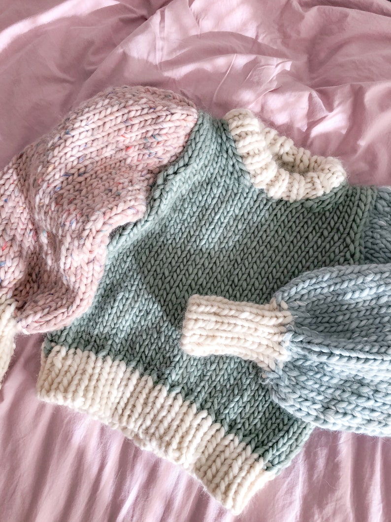 May include: A chunky knit sweater with a color block design. The sweater has a pink and white speckled sleeve, a green body, and a blue sleeve. The sweater is laying on a pink surface.